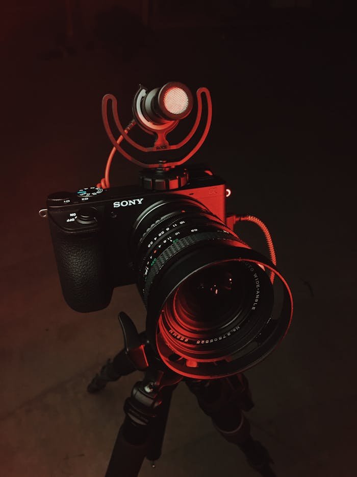 A Sony camera with lens and microphone mounted on a tripod, illuminated by red lighting.